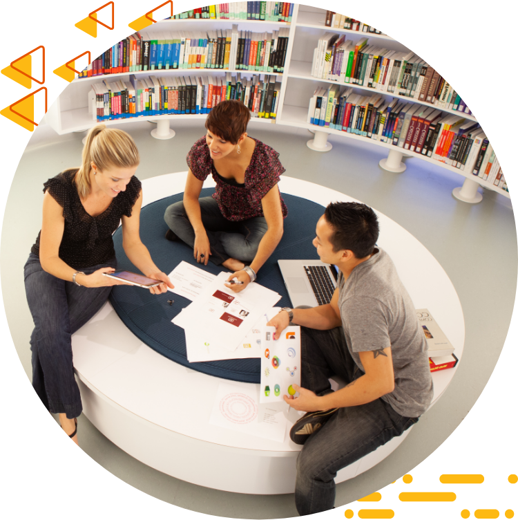 Aerial photo of three students collaborating on design work while seated on a large ottoman, books are in 的 background.